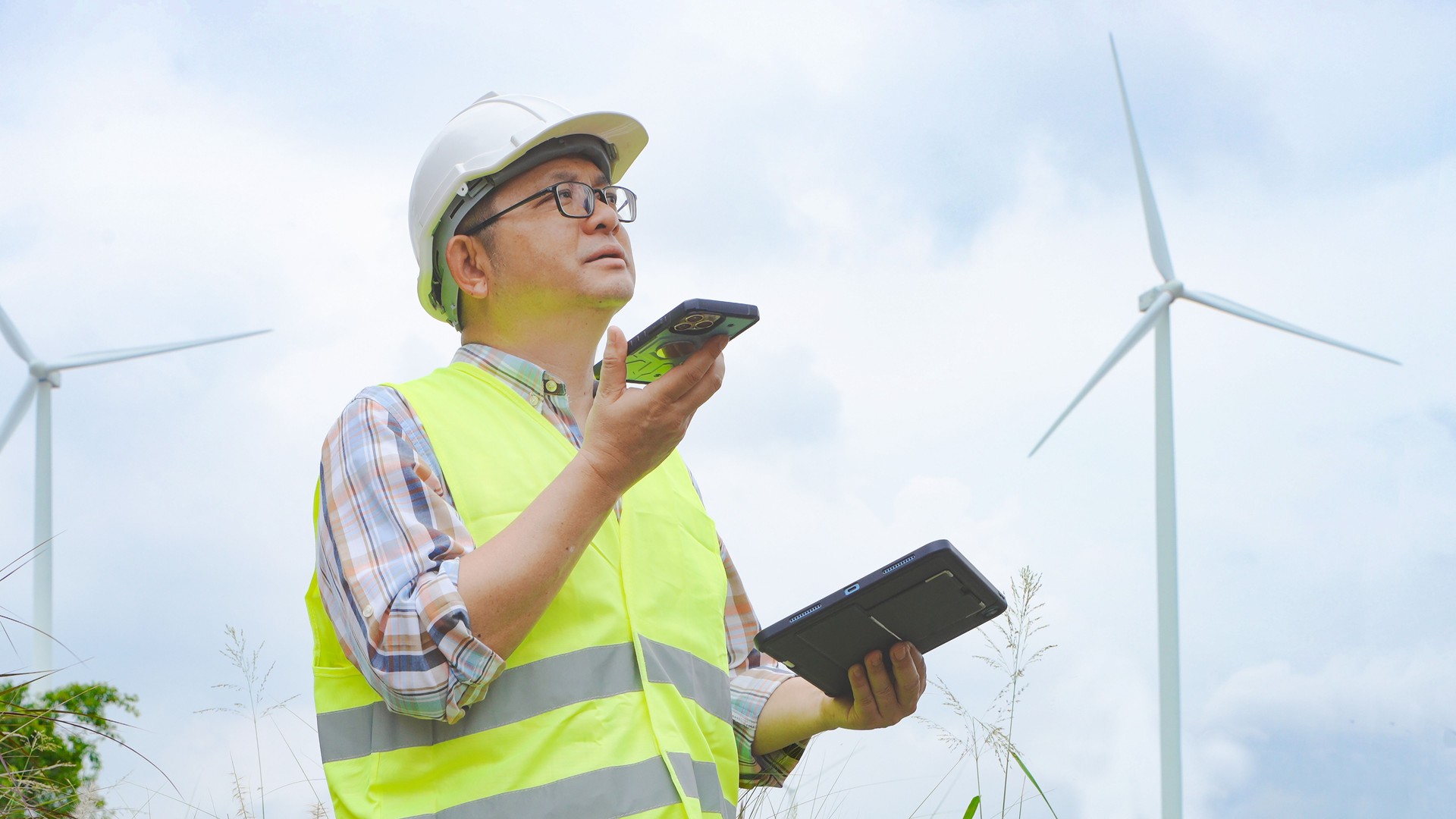 El ingeniero se comunica a través de un teléfono inteligente mientras sostiene la tableta durante la inspección de molinos de viento en un sitio de energía renovable
